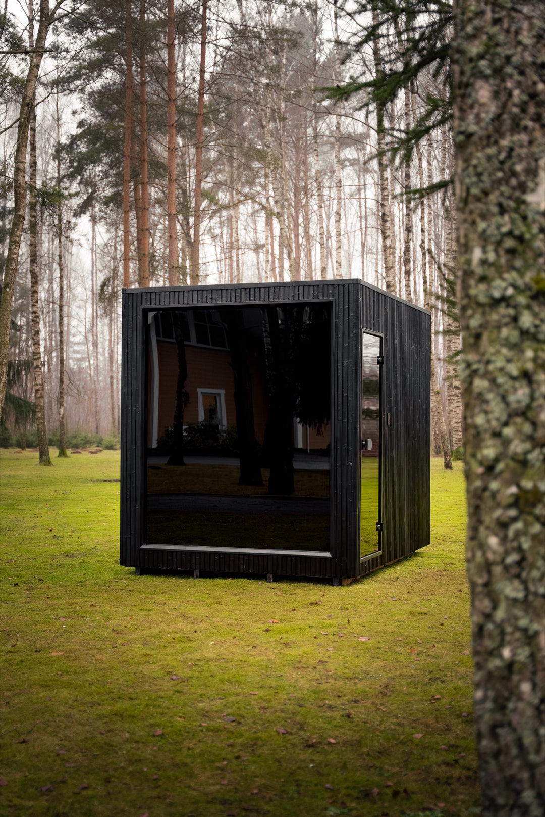 Sauna ogrodowa VALO L dla 6–8 osób – czarna elewacja, pełne przeszklenie frontu i szklane drzwi, wśród brzóz