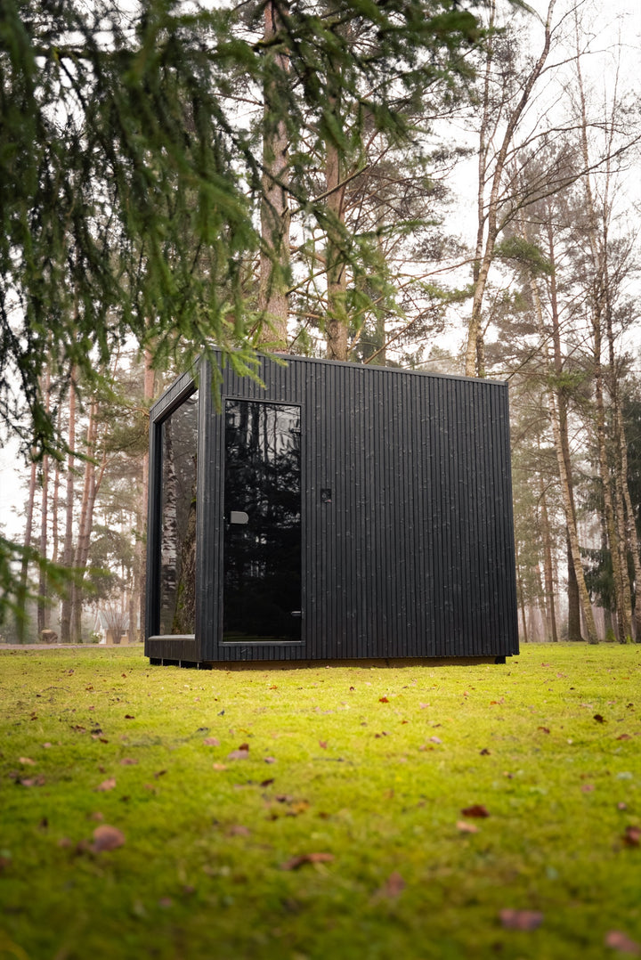 VALO L large cube – garden sauna with glass door under spruce branches, black board cladding
