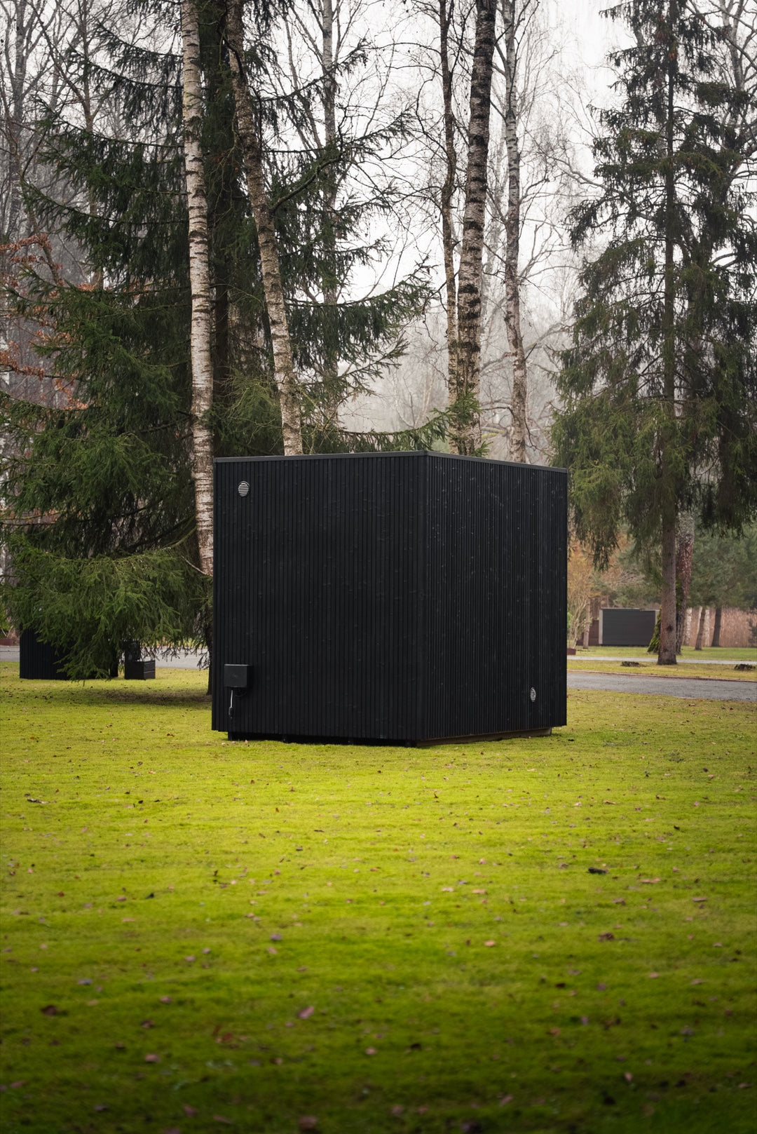 Rear wall of the VALO L garden sauna – solid black cladding without glazing on a lawn between spruces and birches