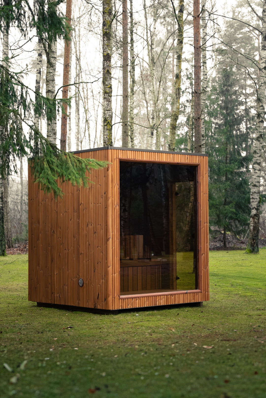 Sauna ogrodowa VALO S – kubik z drewna w ciepłym brązowym odcieniu i pełnym przeszkleniem frontu, na trawniku w lesie